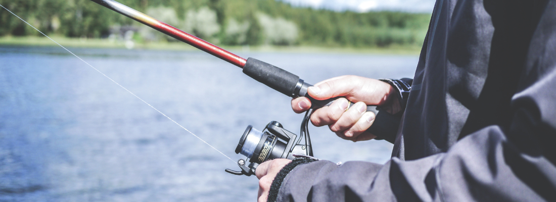 Närbild på en persons händer som håller i ett fiskespö. I bakgrunden syns en sjö.