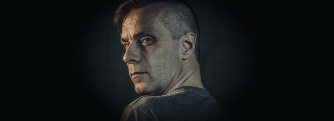 Peter tittar över axeln in i kamera med en mystisk blick. Fotot är mörkt men effektfullt ljussatt.